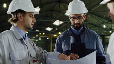 close-up view of three engineers wearing helmets and holding blueprint while talking in a factory