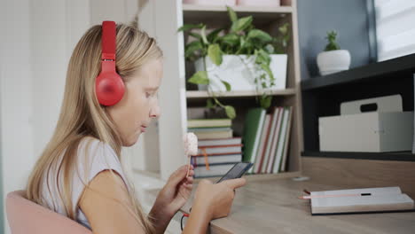 Una-Adolescente-Con-Auriculares-Usa-Un-Teléfono-Inteligente,-Come-Helado-En-Su-Habitación