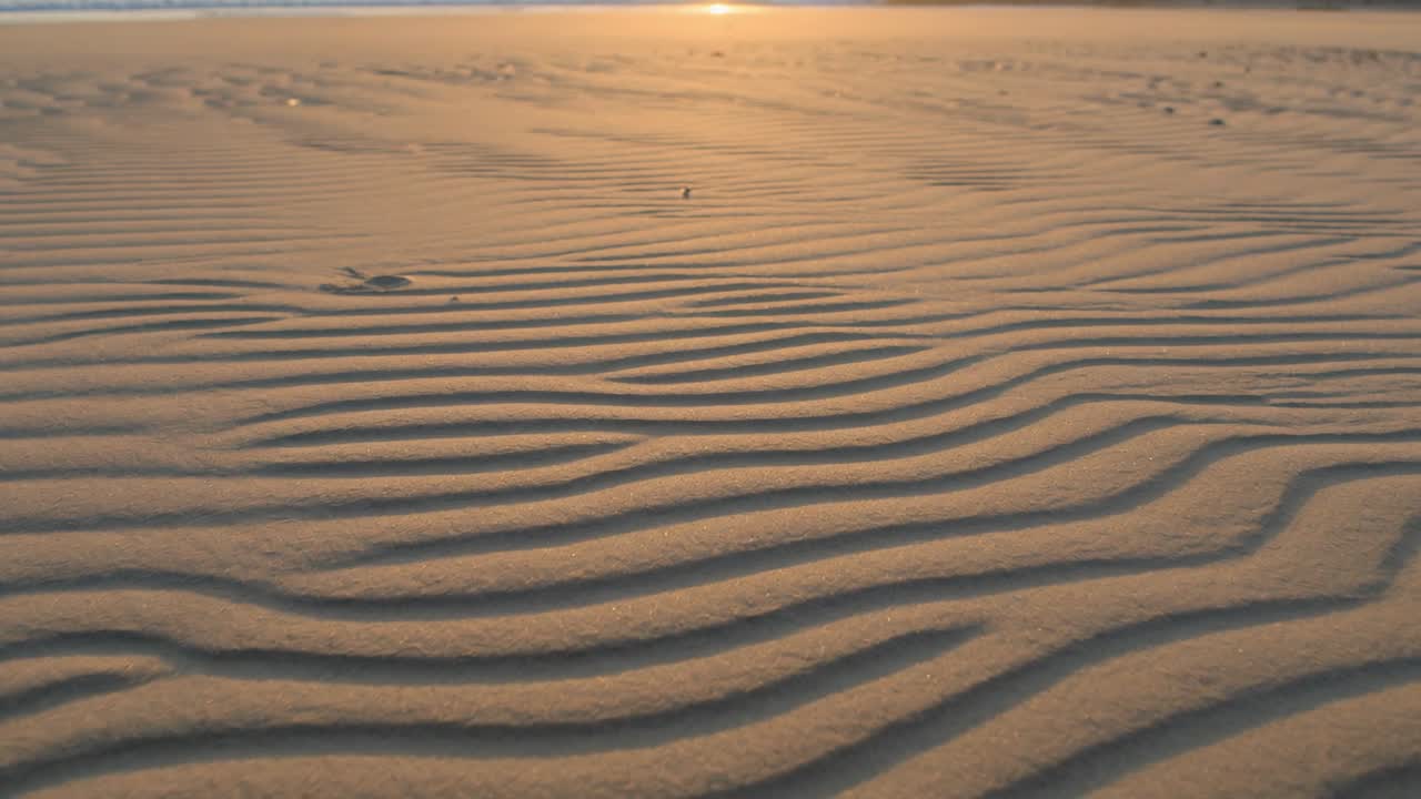 Mostrando increspature di sabbia formate dal vento mentre la telecamera si sposta verso la costa sotto il sole basso, esponendo un oggetto scuro.