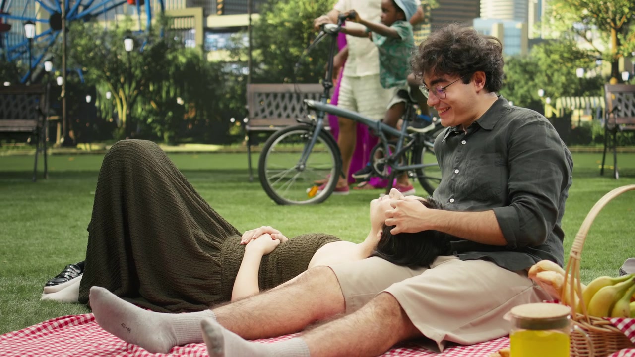 Man gently holds and caresses young woman resting on his lap on picnic blanket,