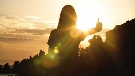 young girl with long hair silhouetted by rocky landscape gives thumbs up with bright white sun and glare shining in background sky, static