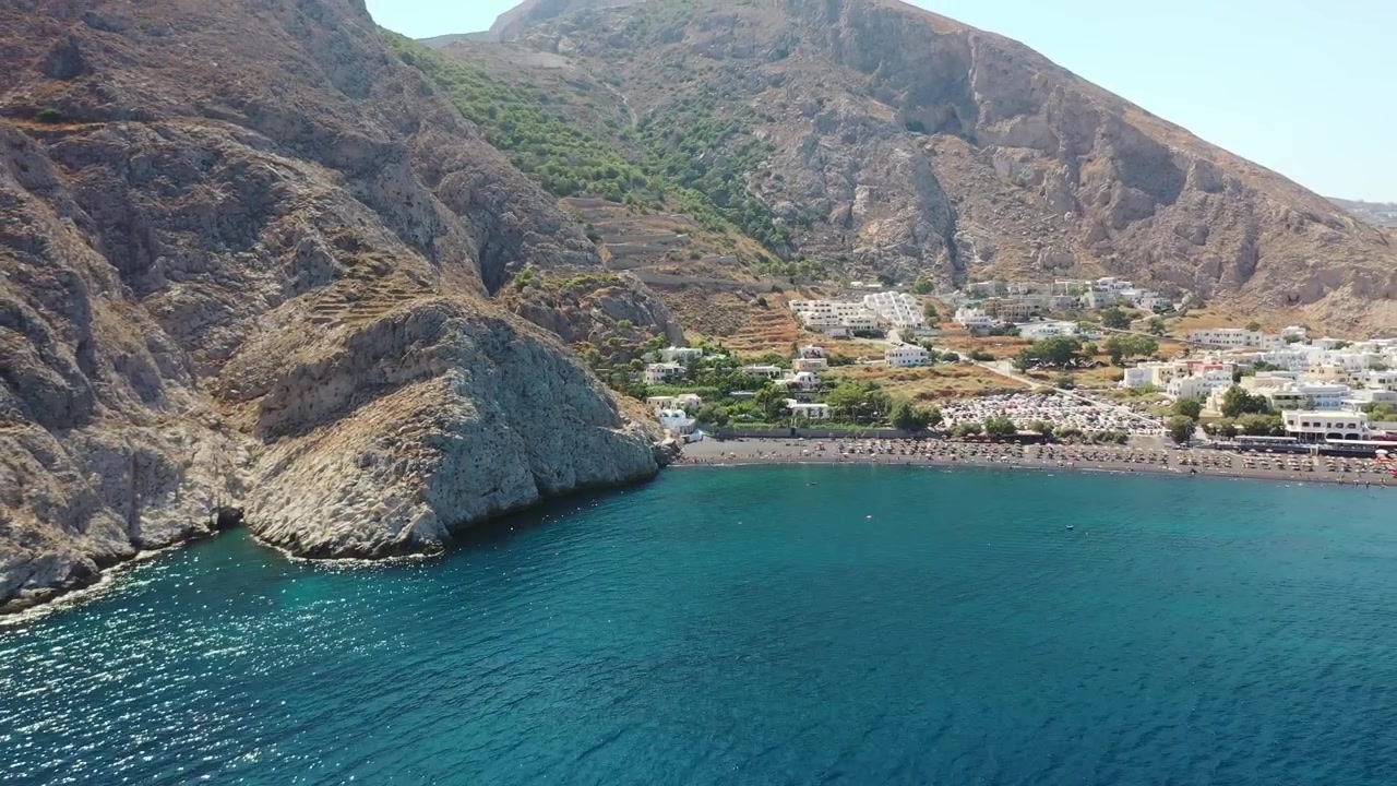Mesa Vouno'nun derin mavi Ege Denizi'ne inen devasa kayalık uçurumlarını yakalayan çarpıcı drone çekimi. Parlak bir yaz gökyüzünün altında Perissa sahil kasabasının siyah kumlu kıyı şeridini gösteriyor.