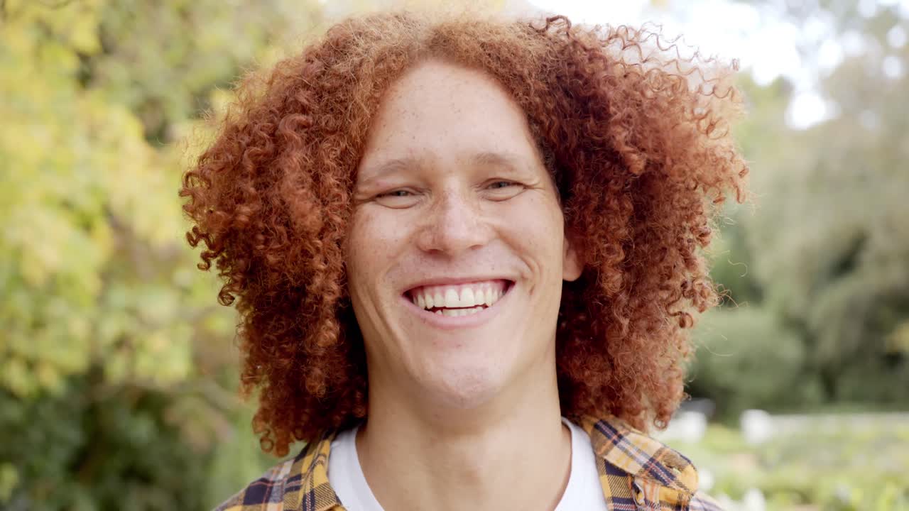 retrato de un feliz hombre biracial con cabello rojo rizado y pecas sonriendo en el jardín, cámara lenta
