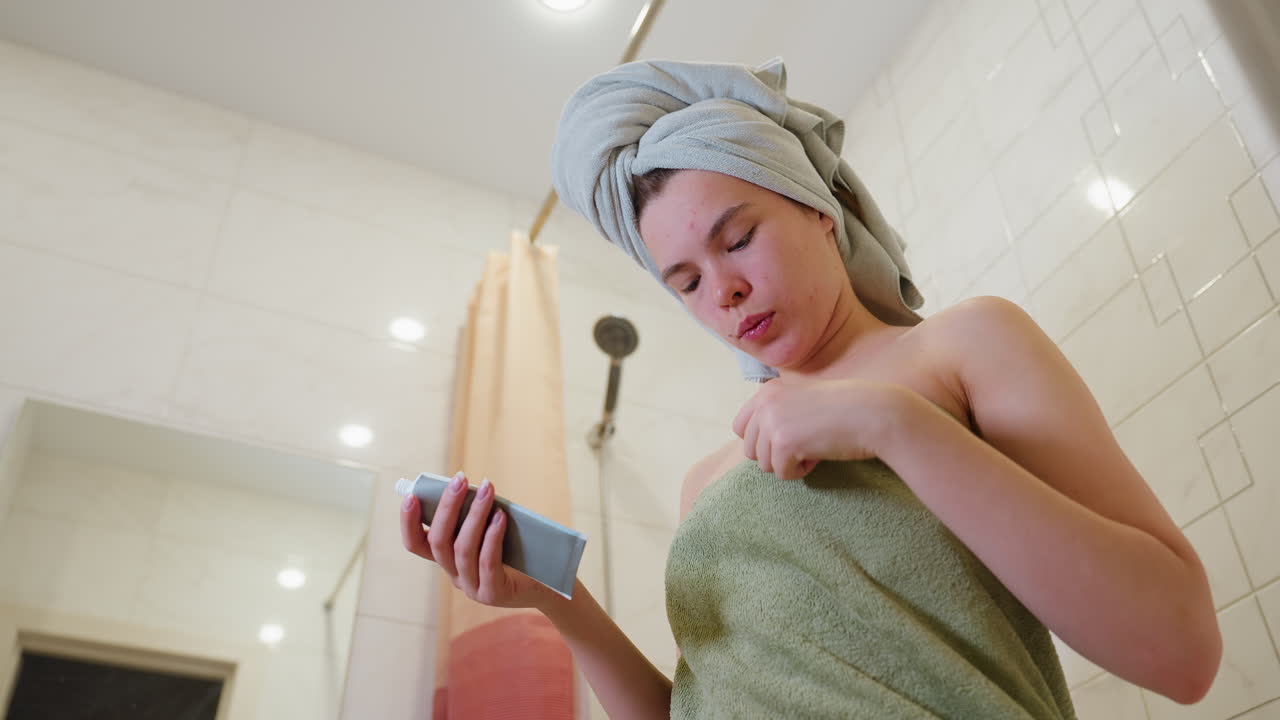 Close view of lady adjusting towel to chest while holding cream tube in bathroom after shower, preparing for skincare routine, showing relaxed mood and natural moment of self-care