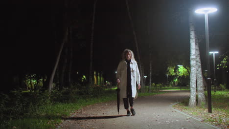 young lady in long white coat walking slowly along peaceful park pathway illuminated by glowing streetlights, holding umbrella, surrounded by trees and autumn leaves in serene