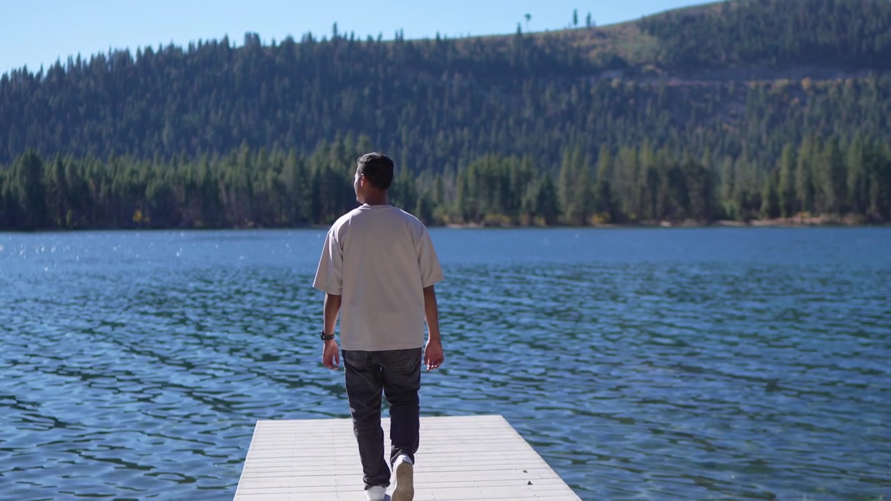 Cinematic rear view shot of a male walking on a lakeside jetty in slow motion with peaceful natural surroundings and scenic blue water.