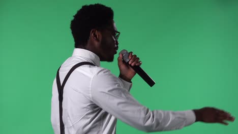 retrato de un joven afroamericano cantando felizmente en el micrófono y gestando con las manos. hombre negro con corbata y gafas en camisa blanca posando en pantalla verde en el estudio. vista lateral.