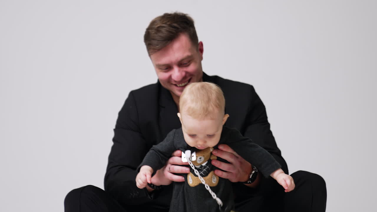 Beautiful fair-haired baby with a teddy on his body suit and attached pacifier. Smiling father holding his son under arms. White backdrop.