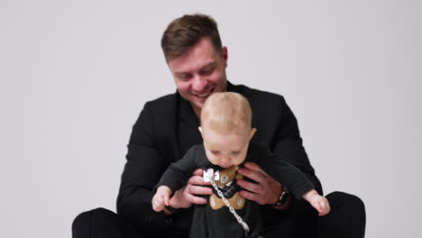 beautiful fair-haired baby with a teddy on his body suit and attached pacifier. smiling father holding his son under arms. white backdrop.