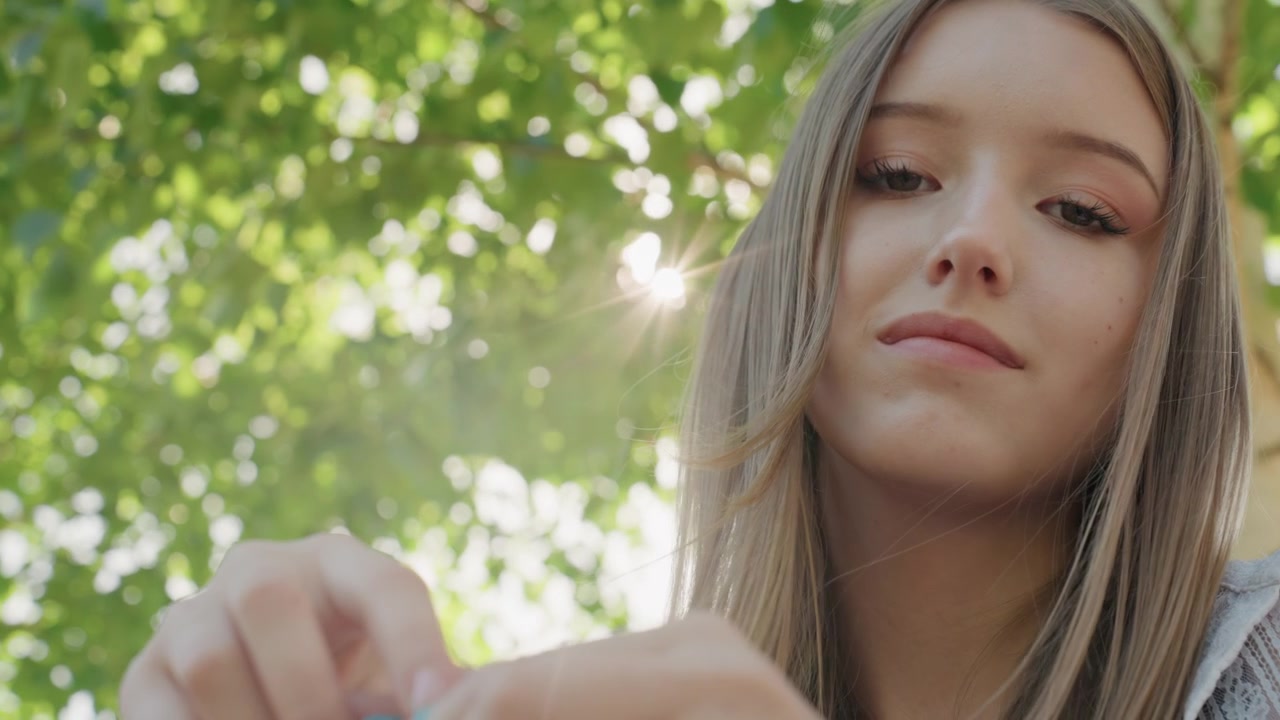 Primo piano di una giovane donna che lavora con filato blu, le mani che lavorano attivamente all'uncinetto o a maglia in un ambiente esterno. La scena presenta una luce naturale soffusa e uno sfondo di fogliame verde.