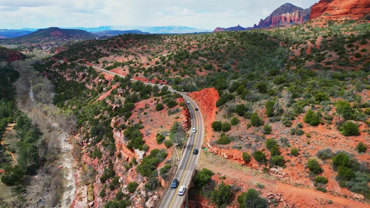 An aerial view captures cars crossing Midgley Bridge in Sedona, Arizona. The bridge spans a river valley, framed by iconic red rock formations and vibrant desert greenery under a clear sky