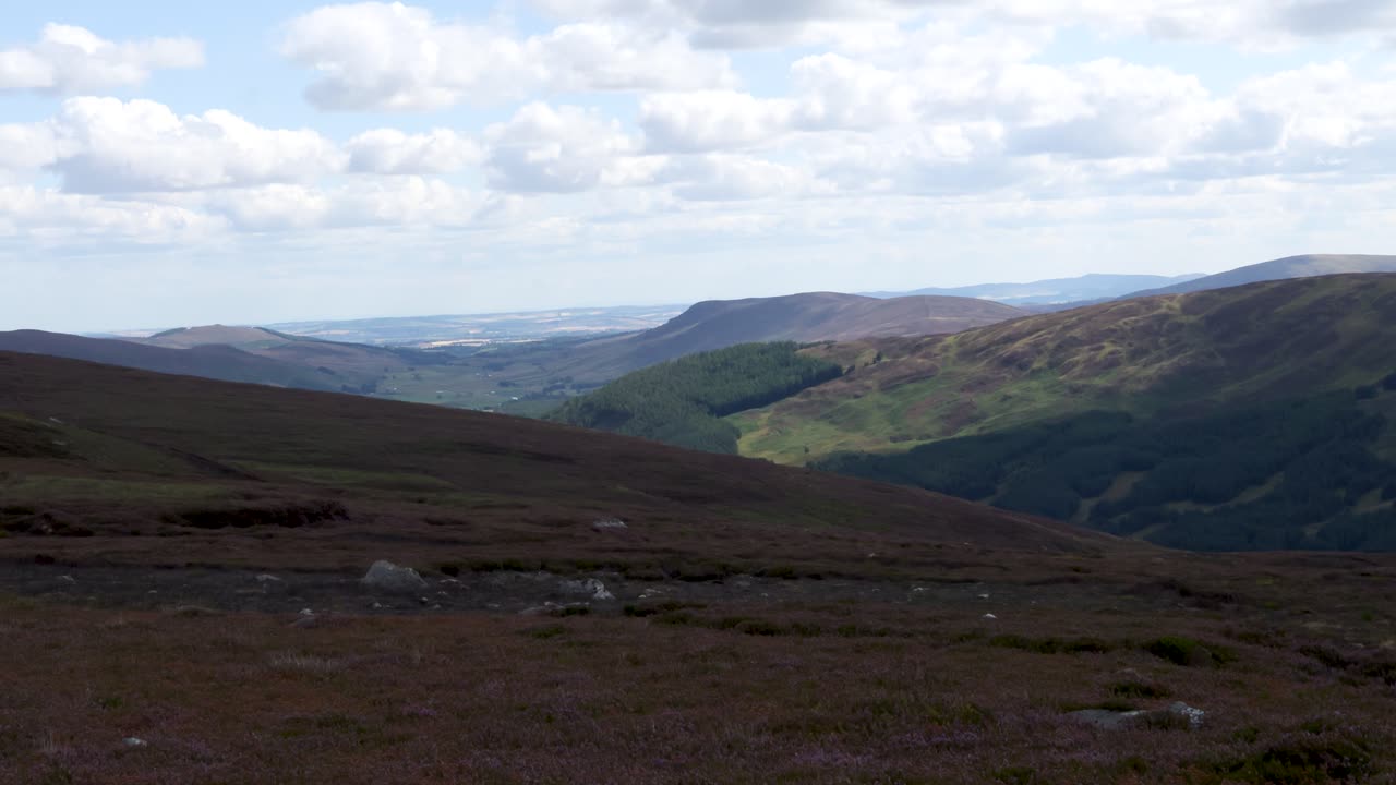 Wide daytime pan reveals rolling heather moorland, green valleys, and distant hills under cloudy sky