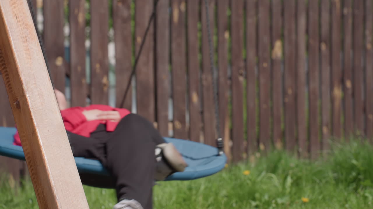Close up of lady resting on swing with sunglasses on, eyes closed, as warm sunlight reflects on her face, she lies peacefully surrounded by tall green grass near wooden fence in calm outdoor setting
