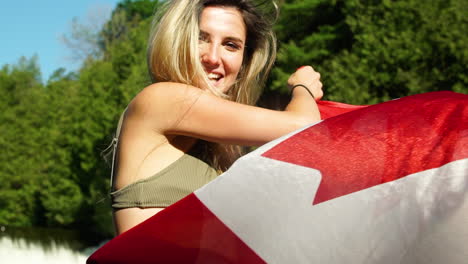 confident beautiful young woman in bikini waves a canadian flag at the camera while posing in a river