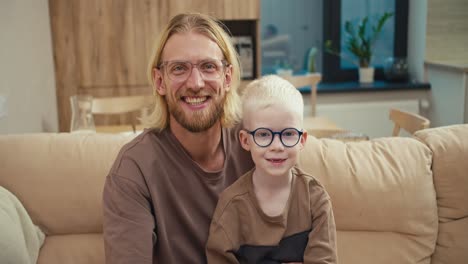 Portrait-of-a-happy-blond-man-in-glasses-and-his-little-albino-son-in-blue-glasses-who-are-sitting-on-a-cream-sofa-in-a-modern-apartment.-A-little-albino-boy-with-white-hair-wearing-blue-glasses-sits-and-rejoices-with-his-father-in-a-modern-apartment-in-front-of-school-in-the-morning