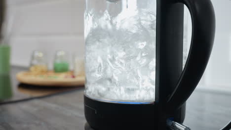 close up of water boiling rapidly in transparent jar with bubbles forming and rising, accompanied by a soft blue light, background features various kitchen equipment and blurred objects
