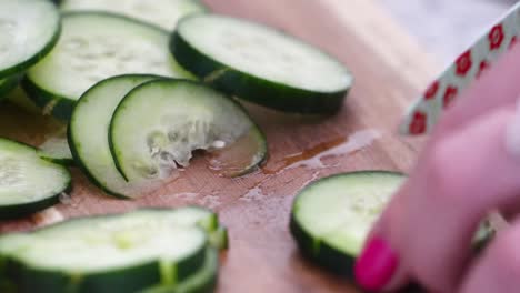 woman chopping cucumber slices into cubes and chunks, close up on wooden cutting board with small knife decorated with flowers, hands with pink nail polish