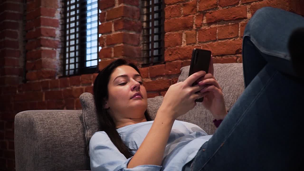 la joven está tumbada en el sofá de la sala de estar con una pared de ladrillo rojo y está escribiendo un mensaje con su teléfono inteligente, tomada desde un lado, en primer plano