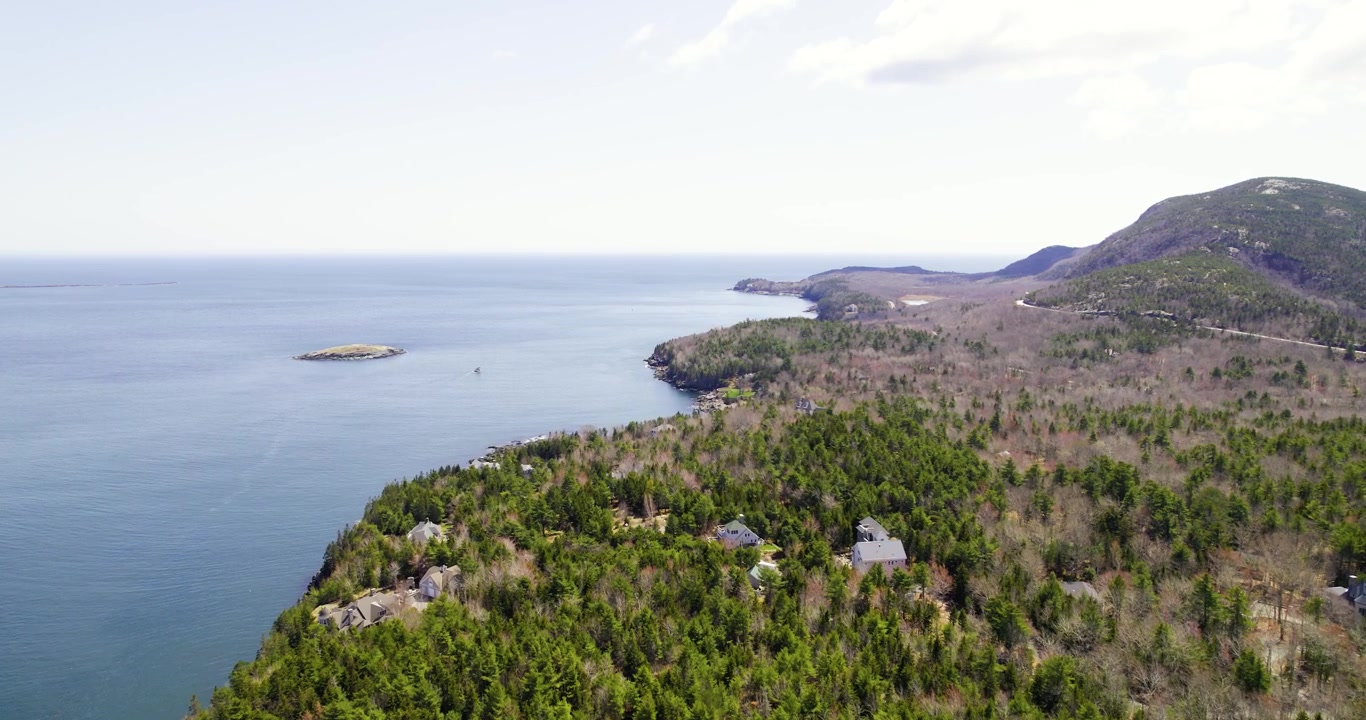Penerbangan drone yang mulus mengikuti pemandangan laut alami Maine berupa air biru, bebatuan, dan lahan berhutan di bawah sinar matahari yang cerah