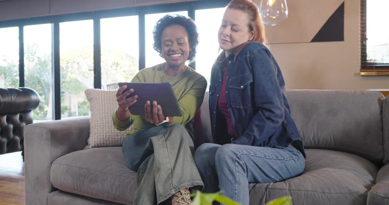 Diverse female friends reacting to tablet content, passing device on sofa and discussing details