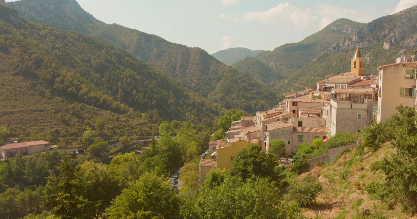 Veduta dal fianco della collina di Sainte-Agnès, uno dei borghi più belli della Francia, con edifici in pietra e il campanile della chiesa che si ergono dai pendii montuosi boscosi. Ideale per contenuti di viaggio e patrimonio culturale.