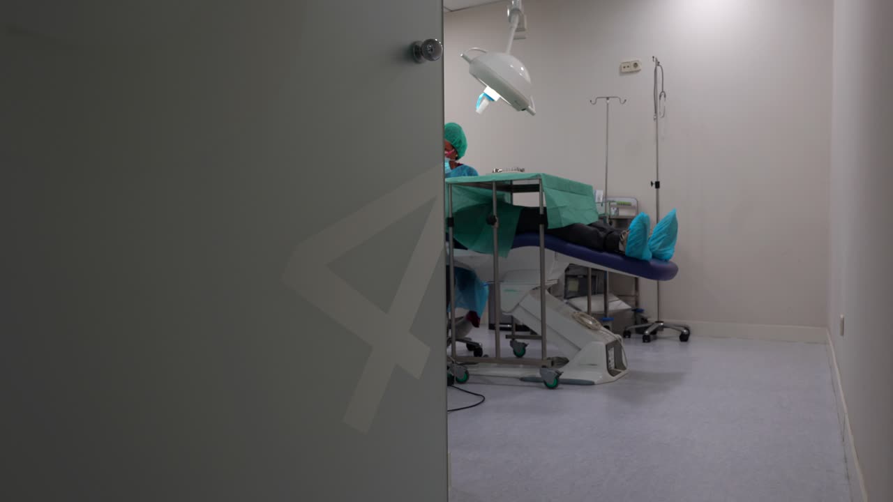 Sliding shot of dentist treating patient on chair as assistant enters clinic room