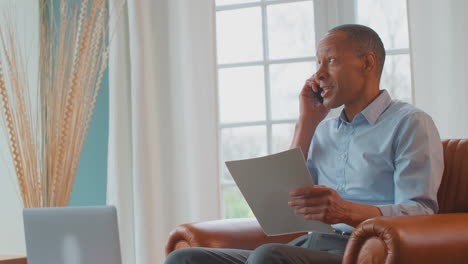 mature man or businessman with mobile phone working from home in armchair looking through paperwork