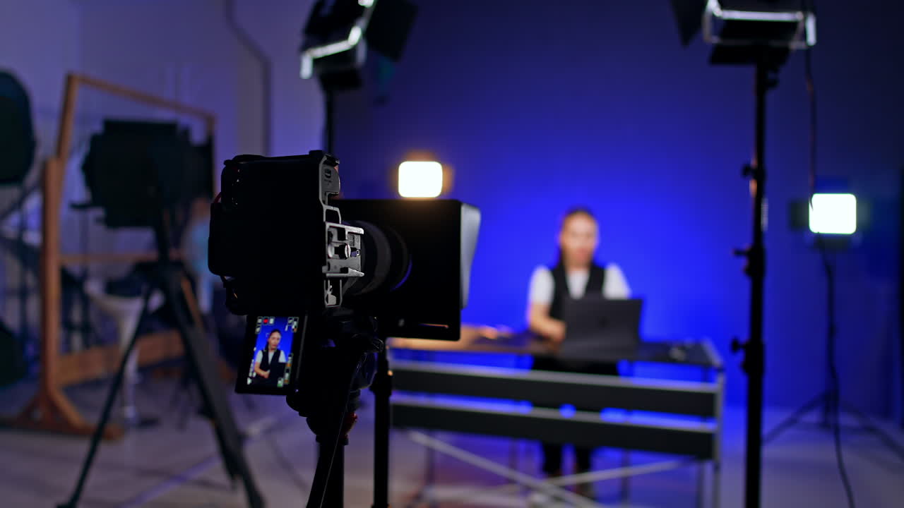 Camera on tripod records a woman in studio. Woman works at her laptop at blurred backdrop. Male photographer comes up to desk putting something.