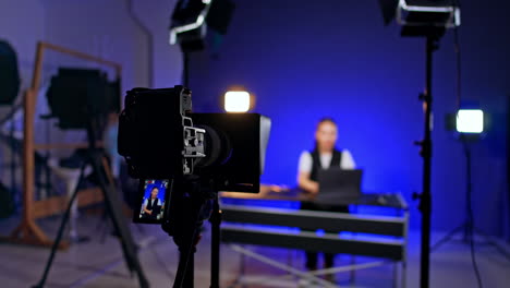camera on tripod records a woman in studio. woman works at her laptop at blurred backdrop. male photographer comes up to desk putting something.