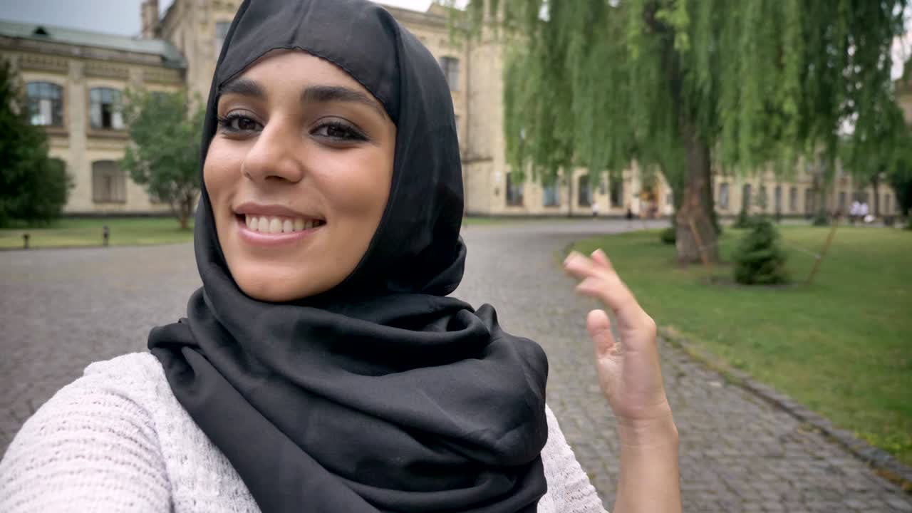 Young happy muslim woman in hijab is making video, saying hey, waving hand in blue weather, communication concept, religious concept, building on background