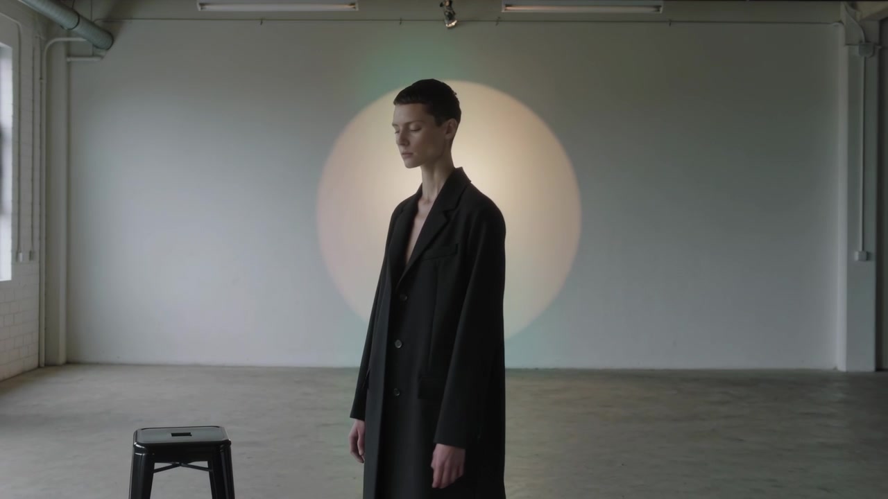 Abbassando lo sguardo, modella in lungo cappotto nero che gira oltre uno sgabello in studio verso una luce circolare per un ritratto