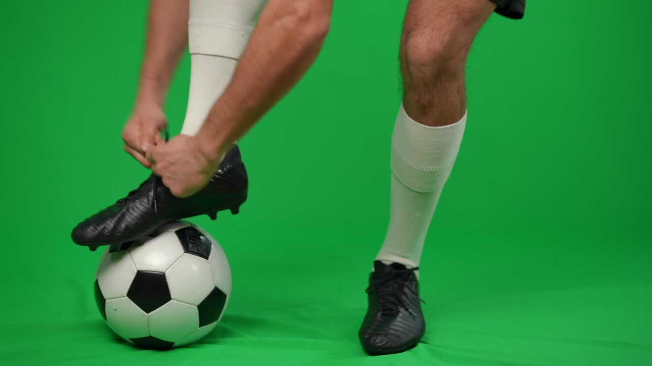 Sportivo caucasico che appoggia il piede sul pallone da calcio allacciandosi le scarpe da ginnastica. Giovane calciatore professionista irriconoscibile su sfondo green screen in chromakey.