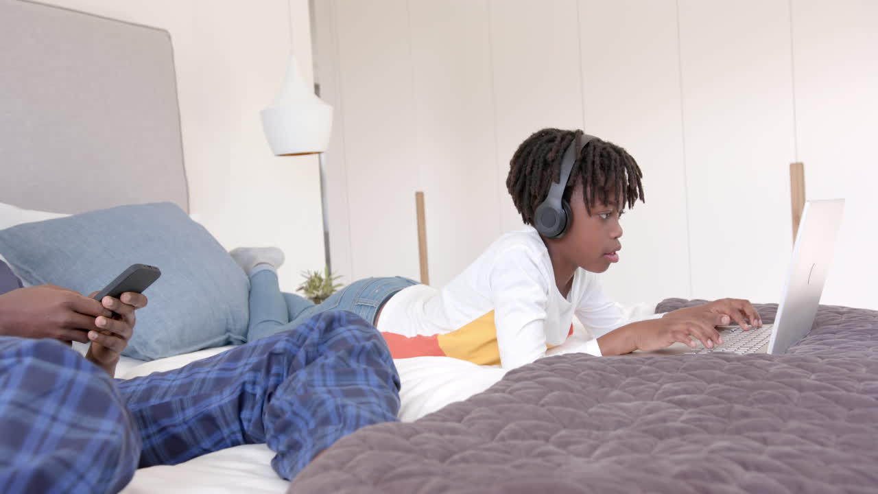 African american father and son lying on bed using laptop and smartphone at home, slow motion