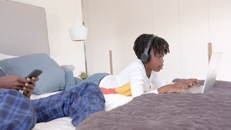 african american father and son lying on bed using laptop and smartphone at home, slow motion