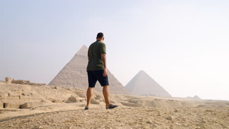 a man walks through desert at giza plateau with pyramids at background in giza, egypt. tracking shot