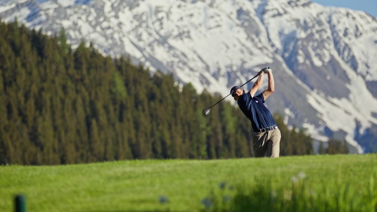 Professional golfer taking a full swing on the fairway at Verbier Golf Club in Switzerland. The snow-covered peak of Le Rogneux rises above the alpine course, creating a spectacular mountain golf sett
