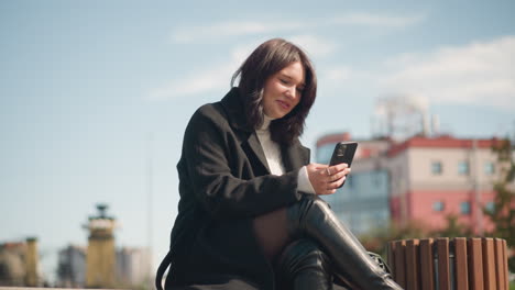 una chica blanca feliz sonriendo mientras usa su teléfono inteligente, vestida con un abrigo negro y un suéter en un parque con las piernas cruzadas, un edificio moderno borroso en el fondo