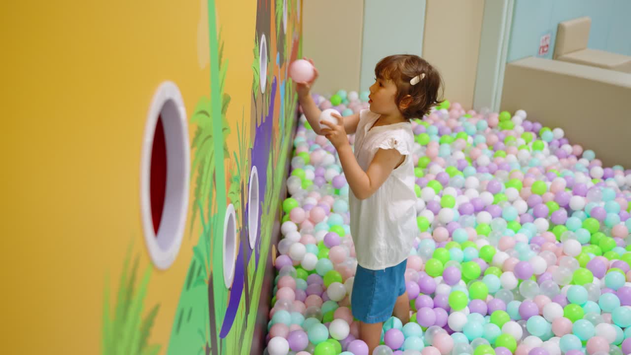 Cute little girl playing and having fun in a colorful ball pit, throwing balls in wall holes at an indoor playground or entertainment center for kids