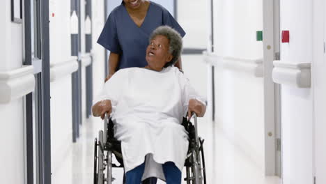 african american female doctor walking with senior woman in wheelchair in hospital, slow motion