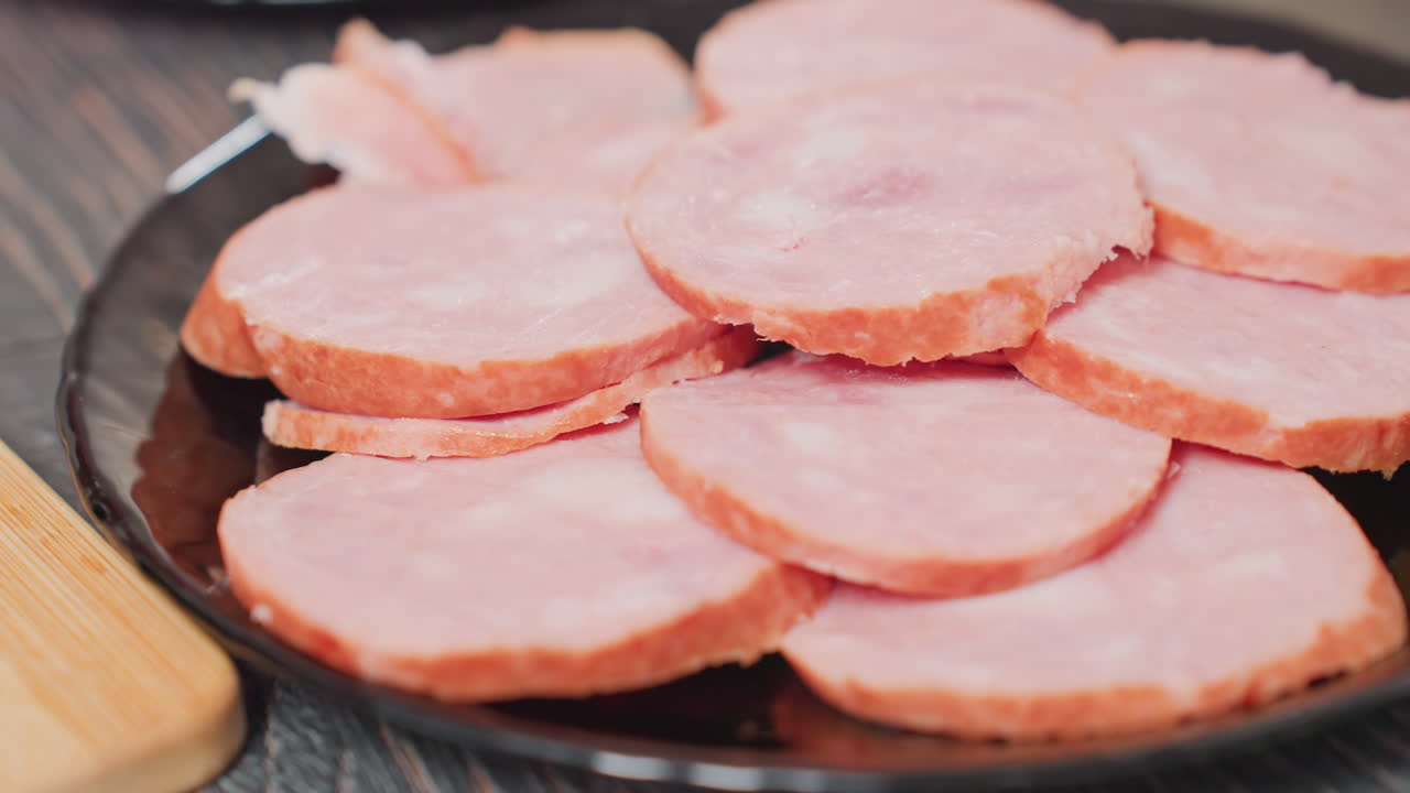 close up of person arranging circular slices of pink hotdog on shiny black plate, overlapping pieces neatly placed for meal prep on dark wooden surface with warm lighting and kitchen ambiance