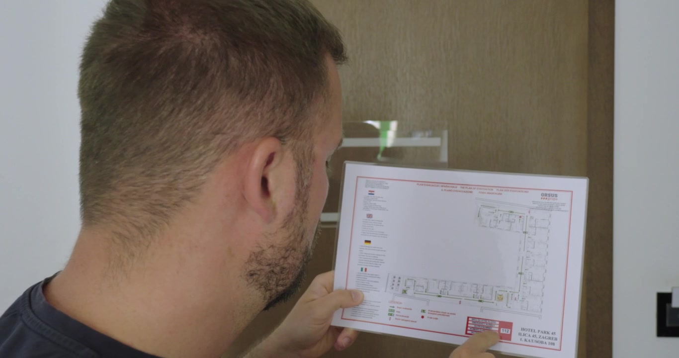 Uomo che legge il piano di evacuazione d'emergenza affisso in una camera d'albergo. Concetto di viaggio sulla sicurezza alberghiera, evacuazione antincendio, preparazione alle emergenze e informazioni per gli ospiti
