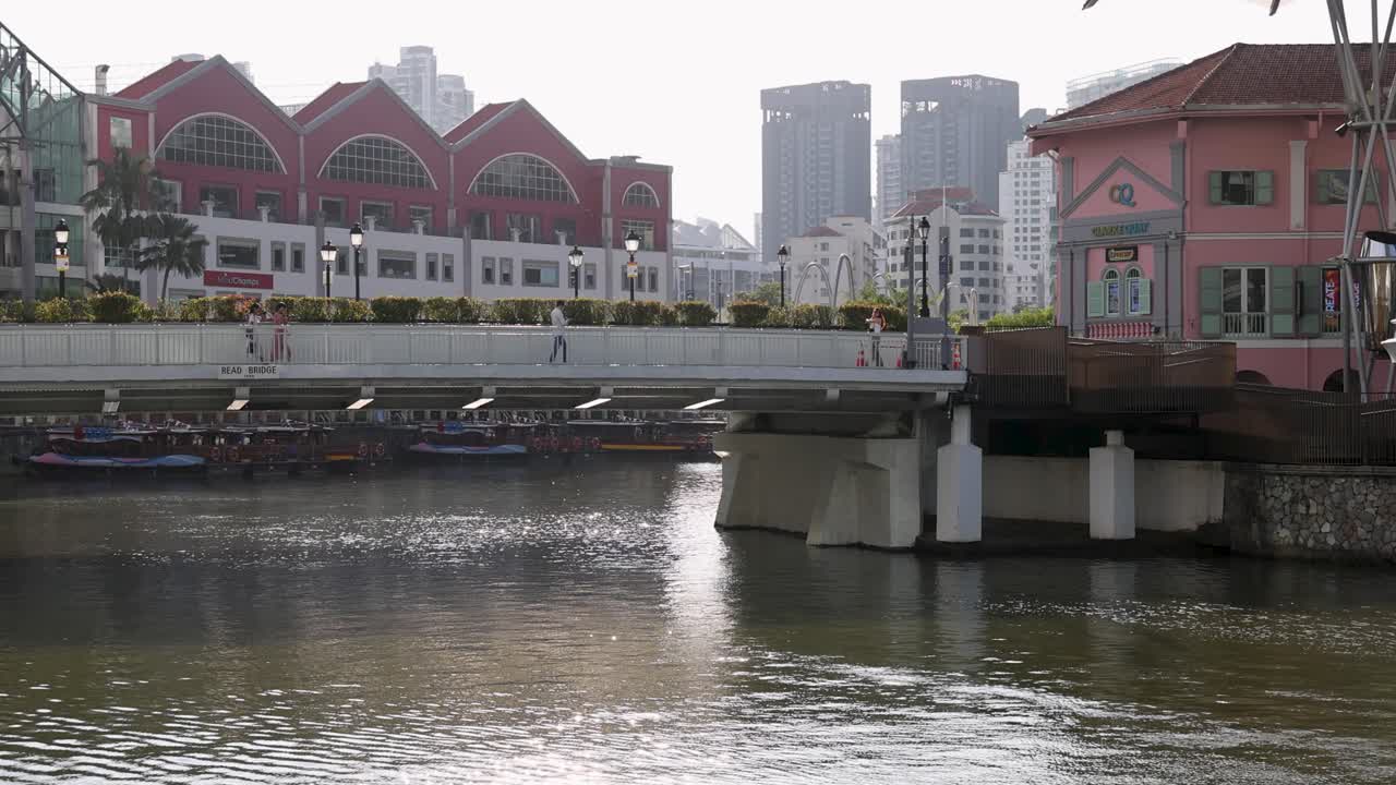 A traditional river cruise boat glides beneath a bridge at Clarke Quay, Singapore, with modern buildings and waterfront architecture in soft daylight. Static wide shot