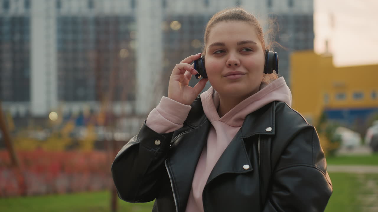 Chubby lady in pink hoodie and black leather jacket gently holds headphones with closed eyes as she enjoys music while breeze softly blows her hair surrounded by blurred urban background