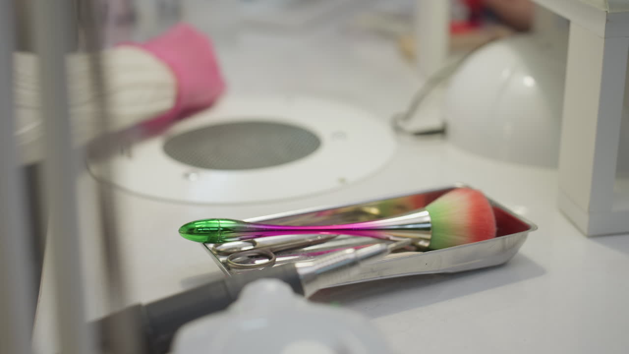 Closeup view of nail technician in pink gloves cleaning work table after manicure, with colorful brush and metal tools arranged in tray beside electric nail drill in clean professional salon setting