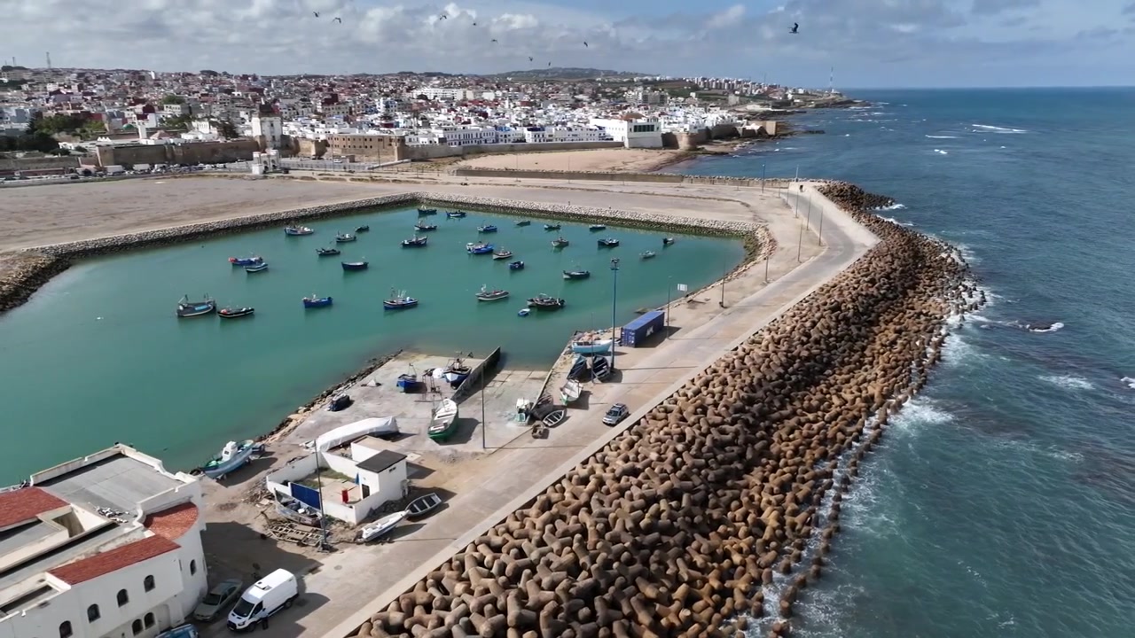 Vista aerea del porto peschereccio di Asilah sulla costa atlantica del Marocco, che mostra un bacino protetto pieno di piccole barche da pesca, lunghe dighe frangiflutti e la storica città costiera oltre.