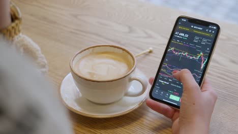 A-woman-checks-the-cryptocurrency-trading-chart-on-her-smartphone-while-drinking-coffee---close-up-shot