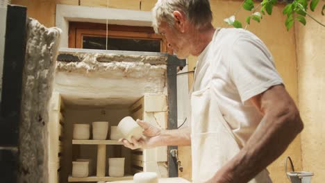 Senior-caucasian-man-wearing-apron-firing-pots-in-kiln-at-pottery-workshop