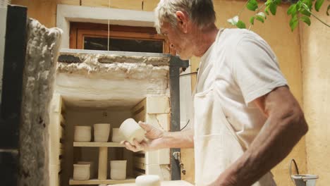 senior caucasian man wearing apron firing pots in kiln at pottery workshop