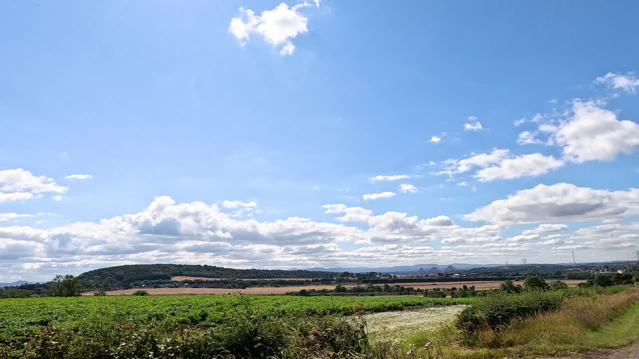Wide panning shot over lush green fields, rolling hills, and blue sky under bright daylight