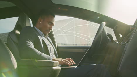 african american man in business suit sitting on drivers seat of modern electric car and typing in